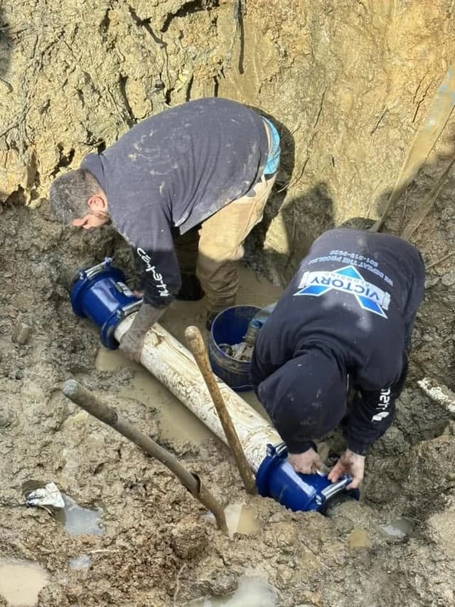 Two Victory plumbers fitting blue couplings onto a water line in the trench